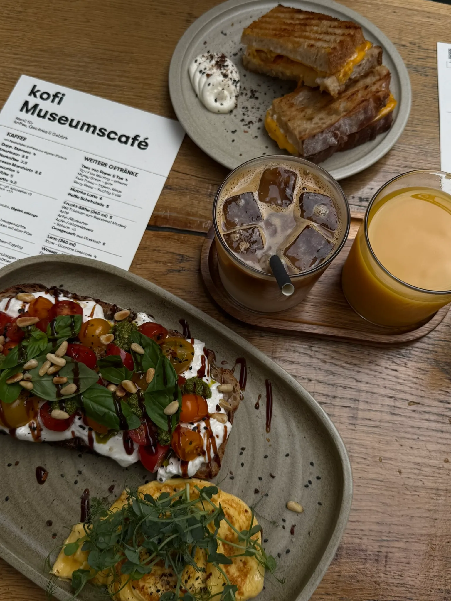 Frühstückstisch im kofi Museumscafé: Tomaten-Burrata-Stulle, getoastetes Sandwich, Iced Latte, Orangensaft und die Speisekarte